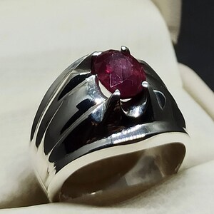 Könnte beinhalten: Ein silberner Ring mit einem tief roten Edelstein, der in der Mitte eingesetzt ist. Der Ring hat ein breites Band mit einem strukturierten Design.