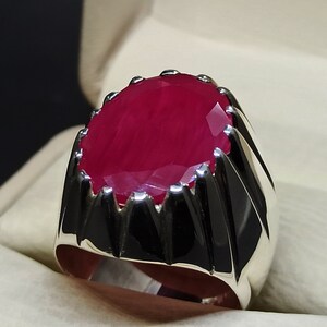 May include: A silver ring with a large, oval-shaped ruby gemstone. The ring has a textured band with a pointed design.