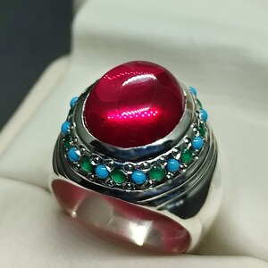 May include: A silver ring featuring a large, oval-shaped, red gemstone set within a band of smaller turquoise and green gemstones.