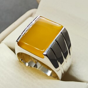 May include: A silver ring with a yellow gemstone set in a square shape. The ring has a textured band with three raised lines.