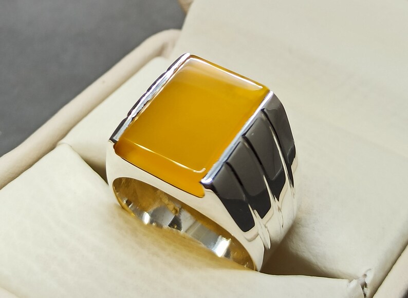 May include: A silver ring with a yellow gemstone set in a square shape. The ring has a textured band with three raised lines.