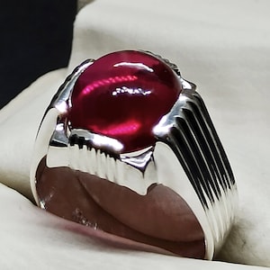 May include: A silver ring with a large, round, red gemstone set in the center. The ring has a textured band with vertical lines.