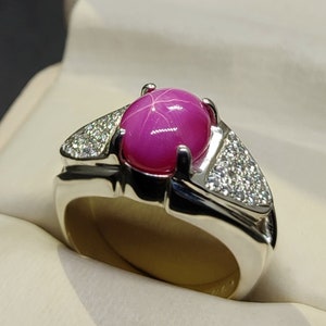 Op de afbeelding: Een zilveren ring met een grote roze ster-robijn edelsteen en kleinere diamanten aan de zijkanten.
