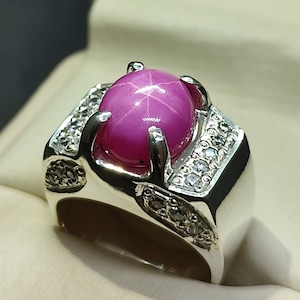 Può includere: Un anello d'argento con una pietra preziosa di rubino stellato rosa e pietre preziose più piccole e chiare intorno alla montatura.