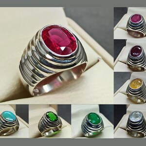 May include: A collection of gemstone rings with a silver band. The rings feature oval-cut gemstones in various colors, including red, yellow, green, and blue. The rings are displayed on a neutral background.