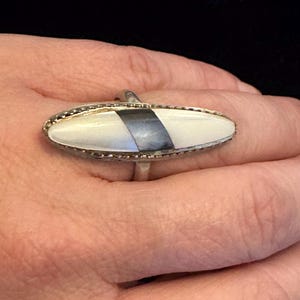 Pode incluir: Um anel de prata com uma pedra oval branca e uma faixa cinza escura. A pedra é engastada numa banda de prata com uma borda decorativa. A joia é um anel.