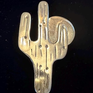 Puede incluir: Un broche de metal plateado con forma de cactus con detalles grabados. El broche presenta una forma de cactus estilizada con múltiples brazos y una sección redondeada. La superficie tiene líneas verticales y pequeñas hendiduras circulares, sobre un fondo oscuro.