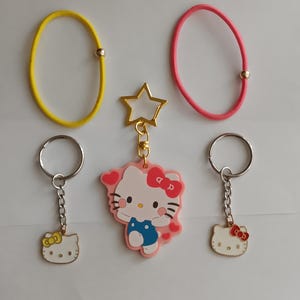 Puede incluir: Una colección de accesorios con temática de Hello Kitty. Incluye dos llaveros con anillos plateados y caras de Hello Kitty, un charm en forma de estrella y dos gomas para el pelo en amarillo y rosa. Los llaveros y el charm tienen detalles dorados.