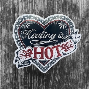 Könnte beinhalten: Ein herzförmiger Aufkleber mit schwarzem Zentrum und rotem Rand. Die Worte "Healing is HOT" sind in Weiß und Rot geschrieben, mit einem Banner-Design. Der Aufkleber hat ein Ketten- und Blumendesign.