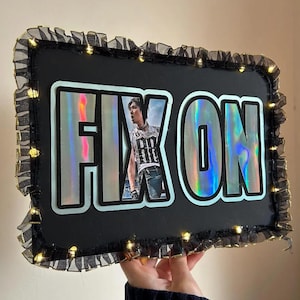 Puede incluir: Un letrero rectangular negro con las palabras "FIX ON" en letras grandes e iridiscentes. El letrero presenta una foto de una persona y está adornado con tela negra con volantes y pequeñas luces blancas cálidas.