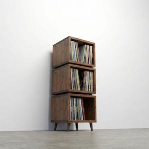 May include: A vertical storage unit composed of three stacked wooden cube compartments, each filled with vinyl records. The unit features a dark brown wood grain finish and is supported by four short, tapered wooden legs at the base.