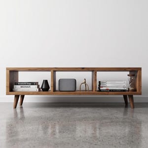 May include: A wooden media unit in a mid-century modern style, featuring three open sections. The unit is crafted from light brown wood and has tapered legs. Books, a black vase, a grey speaker, and decorative items are displayed within.
