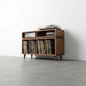 May include: Wooden record player stand with a built-in stereo receiver and a turntable on top. The stand has multiple compartments filled with vinyl records. The unit is made of light brown wood and has a mid-century modern design.