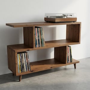 May include: A brown wooden media console with an S-shaped design featuring three open storage levels. The unit stands on four short tapered legs and holds a record player on the top surface, with vinyl records organised in the lower compartments.
