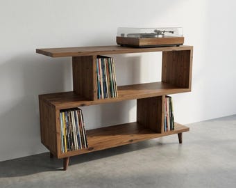 May include: A brown wooden media console with an S-shaped design featuring three open storage levels. The unit stands on four short tapered legs and holds a record player on the top surface, with vinyl records organized in the lower compartments.