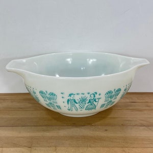 May include: White Pyrex mixing bowl with a turquoise design featuring figures and baskets. The bowl has a wide rim with handles on either side. The bowl is sitting on a wooden surface.
