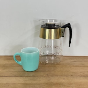 May include: A vintage glass coffee carafe with a gold band and black handle, next to a turquoise mug. The carafe has a decorative brown swirl pattern. The mug is a solid turquoise color.