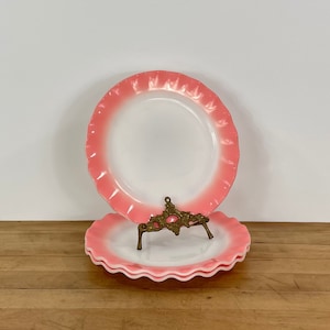 May include: A stack of white and pink plates with a scalloped edge. The plates are displayed on a decorative gold-colored metal stand, with a wooden surface in the foreground and a white background.