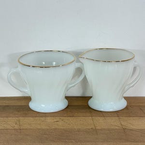 May include: A white milk glass creamer and sugar bowl set. Both pieces have a gold rim and a swirl design. The sugar bowl has two handles. The creamer has a spout for pouring. The set is on a wooden surface.
