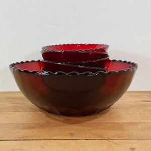 May include: A set of four ruby red glass bowls with scalloped edges. The bowls are nested, with the largest bowl at the bottom and the smallest at the top. The bowls are on a wooden surface.