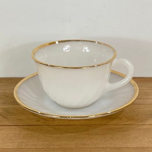 May include: A white teacup and saucer set with a gold rim. The cup has a handle and sits on a matching saucer. The set is made of a glossy material and is suitable for serving tea or coffee. The set is a classic design.