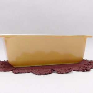 May include: A rectangular, light-orange baking dish with a handle on each end, resting on a dark-red, scalloped-edge mat. The dish has a glossy finish and is set against a white background. This kitchenware is ideal for baking.