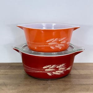 May include: Two vintage Pyrex casserole dishes. The top dish is orange, and the bottom dish is red. Both have a white wheat pattern. The red dish has a clear glass lid. The dishes are on a wooden surface.