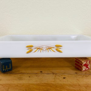 May include: A rectangular white baking dish with a wheat stalk design in gold and brown. The dish is resting on two small blocks on a wooden surface. The dish is approximately 12 inches long.