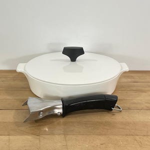 May include: A white enameled cast iron Dutch oven with a black lid handle and a black pot handle. The oval-shaped pot sits on a wooden surface.