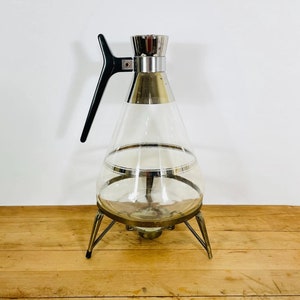May include: A vintage clear glass beverage server with a conical shape, silver trim, and a black handle. The server sits on a brass-toned metal base with three legs. A classic piece for serving drinks.
