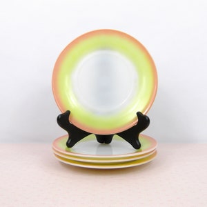 May include: Set of three colorful plates with a white center, transitioning to a light green and then a peach-colored rim. The plates are displayed on a black plate stand, showcasing their design. A stack of two plates is visible below.