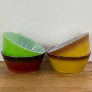 May include: Two sets of vintage Pyrex bowls. One set features a green bowl stacked on a red bowl, the other set has a yellow bowl stacked on a brown bowl. The bowls are on a wooden surface.