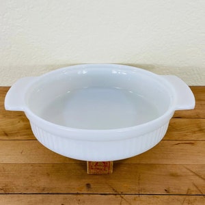 May include: A white, oval-shaped casserole dish with two handles. The dish has a ribbed exterior and a smooth interior. It is sitting on a wooden surface.