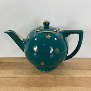 May include: Teal ceramic teapot with gold star accents. The teapot has a rounded body, a curved handle, and a spout. The lid is topped with a gold knob. The teapot is sitting on a wooden surface.