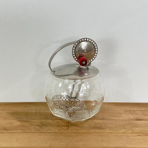 May include: A clear glass butter churn with a silver lid and handle. The churn has a red knob and a metal whisk inside. The glass jar has a hexagonal base and a ribbed design.