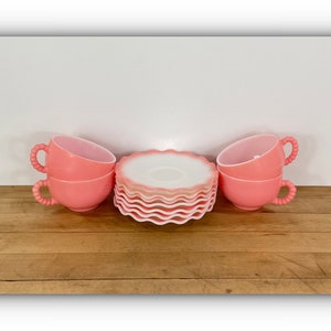 May include: A set of vintage pink teacups and saucers. The cups have a twisted handle design and are stacked in pairs. The saucers have a scalloped edge. The set is arranged on a wooden surface against a white background.
