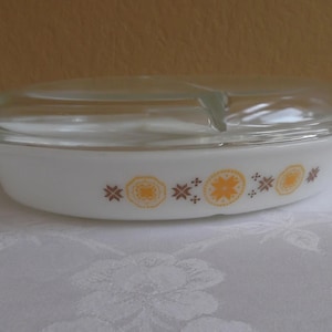May include: A white oval casserole dish with a clear glass lid. The dish has handles and is decorated with a repeating pattern of orange and brown floral designs. A vintage kitchenware item.