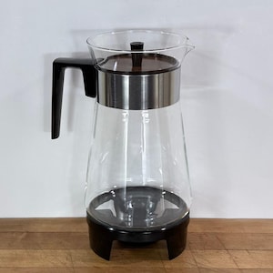 May include: A clear glass pitcher featuring a black plastic handle and a black plastic base. The top includes a stainless steel band and a dark brown wooden lid with a small black knob. The pitcher has a tapered body and a pouring spout.
