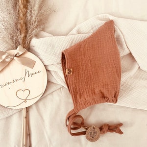 Könnte beinhalten: Ein terracottafarbener Babyhut aus strukturiertem Stoff, mit einem kleinen Holzanhänger mit Regenbogen. Der Hut hat lange Bänder und ein Etikett mit der Aufschrift "Viel Freude damit". Ein Holzschild mit den Worten "nummi Mee" ist ebenfalls im Bild.