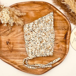 Könnte beinhalten: Ein Baby-Häubchen mit floralem Muster in Creme, Braun und Grün, mit Bindebändern. Das Häubchen liegt auf einem Holztablett mit Trockenblumen und einer Holzscheibe mit der Aufschrift "Mum".