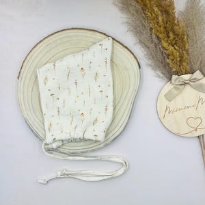 Könnte beinhalten: Ein weißer Babyhut mit zartem Blumenmuster in Braun-, Gelb- und Grüntönen. Der Hut ist aus weichem Stoff gefertigt und hat einen Kordelzug. Er liegt auf einer Holzscheibe.