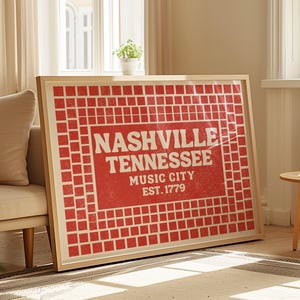 May include: A framed art print featuring the text "Nashville Tennessee Music City Est. 1779" in white on a red grid background. The print is in a light wood frame and rests against a wall near a window.