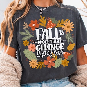 Puede incluir: Camiseta gris oscuro con el texto "FALL is proof that CHANGE is possible" rodeado de ilustraciones florales y de hojas en naranja, amarillo y verde. La camiseta se combina con pantalones cortos vaqueros.