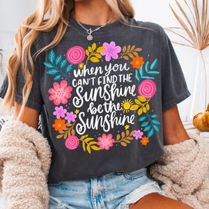 May include: Dark gray t-shirt with the text "when you can't find the sunshine be the sunshine" surrounded by colorful flowers and leaves. The shirt is a relaxed fit and is paired with denim shorts.