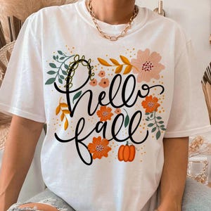 May include: White t-shirt with a floral design and the words "Hello Fall" in black script. The design includes orange flowers, a pumpkin, and green and yellow leaves, perfect for autumn.