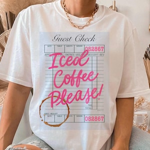 以下が含まれることがあります： 「アイスコーヒーください！」と書かれたレストランのチェックのピンクのグラフィックが描かれた白いTシャツ。コーヒーの茶色のシミがあります。