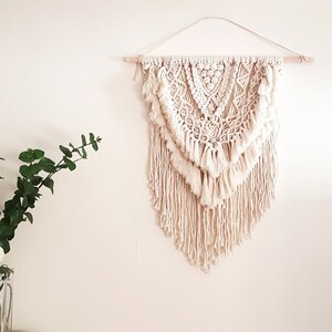 May include: A white macrame wall hanging with a geometric pattern and fringe. The hanging is made of natural fibres and has a bohemian style.