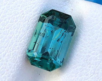 May include: A rectangular-cut gemstone with a teal and green color gradient. The gem has a faceted surface, reflecting light. The gemstone is set against a white, textured background.
