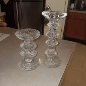 Puede incluir: Dos candelabros de vidrio transparente con superficies texturizadas. Los candelabros tienen un diseño apilado con una base ancha y una parte superior circular. Los candelabros están sobre una superficie de color claro.