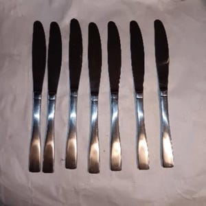 May include: A set of seven silver-toned stainless steel dinner knives arranged in a row. Each knife features a smooth, reflective handle and a serrated blade edge. The knives are positioned vertically on a plain, light-colored surface.